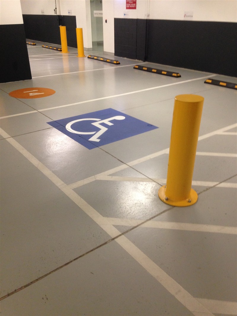 Yellow steel bollard beside an accessible bay, with wheel stops along the bays behind